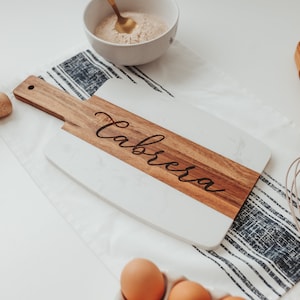 May include: A white and wood cutting board with the name "Cabrera" engraved on the wood portion. The board is on a striped fabric with a whisk and other kitchen items in the background.