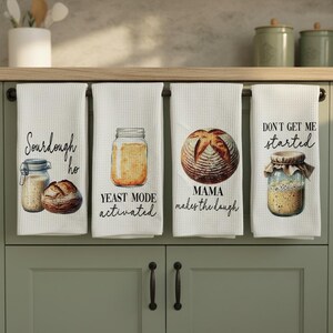 May include: Four white kitchen towels with bread-themed designs and text. One towel says "Sourdough ho" with a jar and bread illustration. Another says "Yeast mode activated" with a jar. The third says "Mama makes the dough" with bread. The last says "Don't get me started" with a jar.