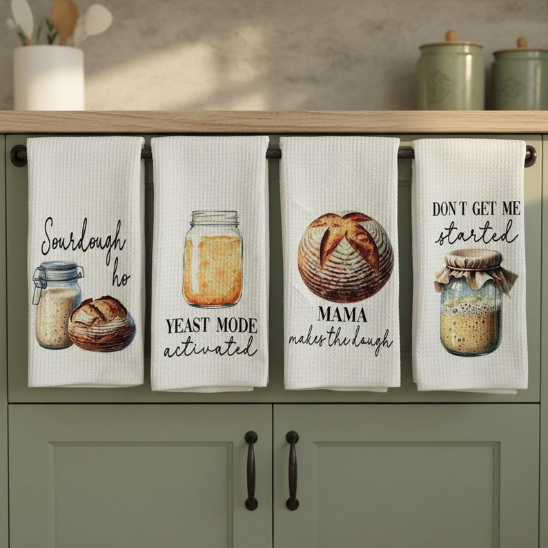 May include: Four white kitchen towels with bread-themed designs and text. One towel says "Sourdough ho" with a jar and bread illustration. Another says "Yeast mode activated" with a jar. The third says "Mama makes the dough" with bread. The last says "Don't get me started" with a jar.