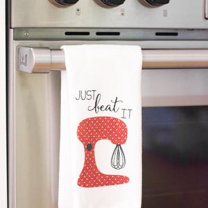 May include: White kitchen towel with a red and white polka dot stand mixer illustration and the text "Just beat it".
