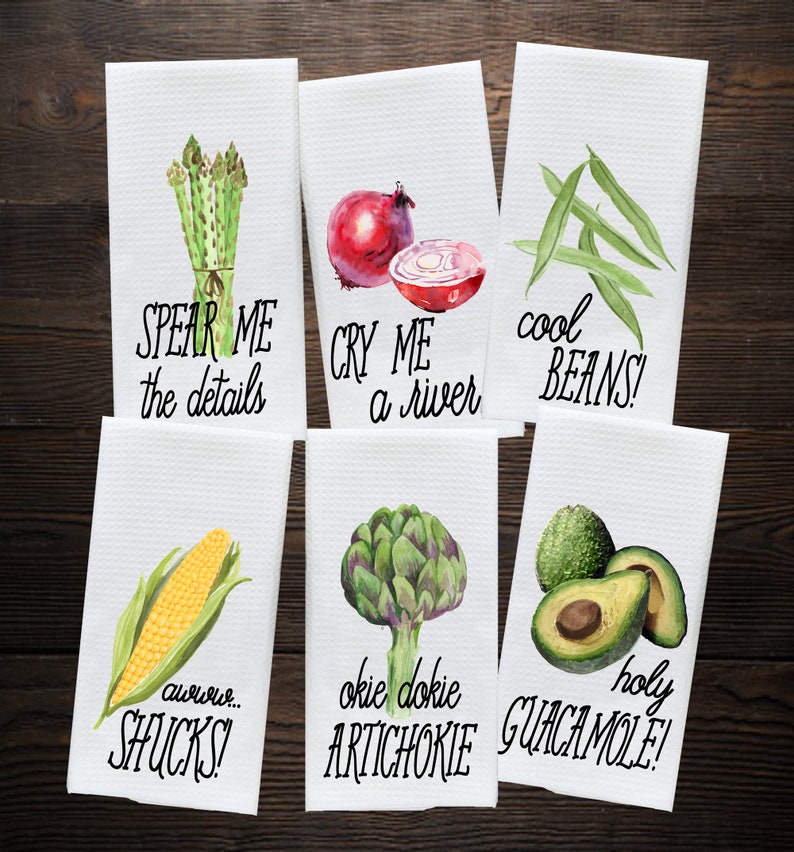 K&ouml;nnte beinhalten: Sechs wei&szlig;e K&uuml;chent&uuml;cher mit Aquarell-Illustrationen von Gem&uuml;se und Wortspielen. Die Handt&uuml;cher zeigen Spargel, Zwiebeln, gr&uuml;ne Bohnen, Mais, Artischocken und Avocados. Der Text auf den Handt&uuml;chern lautet: "Spear Me the details", "Cry Me a River", "Cool Beans", "Aw, Shucks", "Okie Dokie Artichoke", und "Holy Guacamole!"