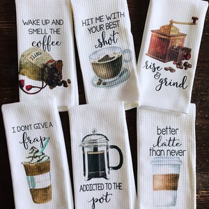 May include: Set of six white kitchen towels with coffee-themed designs and sayings. The towels feature illustrations of coffee beans, coffee cups, a coffee grinder, and a French press. The sayings on the towels include "Wake up and smell the coffee", "Hit me with your best shot", "Rise and grind", "I don't give a frap", "Addicted to the pot", and "Better latte than never".