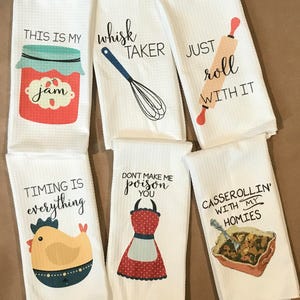 May include: Set of six white kitchen towels with humorous sayings and illustrations. The towels feature sayings like "This is my jam", "Whisk taker", "Just roll with it", "Timing is everything", "Don't make me poison you", and "Casserolin' with my homies".