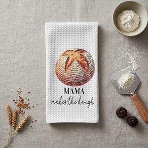 May include: White waffle-weave kitchen towel with a printed image of a loaf of bread. The text "MAMA makes the dough" is printed below the bread image. Wheat stalks, a bowl of cream, and figs are also visible.