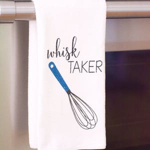 May include: White kitchen towel with a blue and black whisk illustration and the text "whisk taker" in black.
