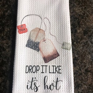 May include: White tea towel with a brown and black tea bag illustration and the text "DROP IT LIKE IT'S HOT".