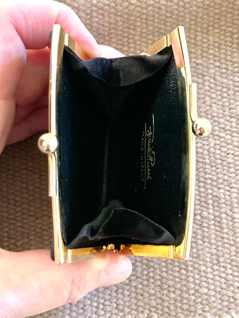 May include: A gold-toned metal coin purse with a black velvet interior. The purse is open and the interior is visible. The purse has the text "Furla" and "Made in Italy" printed on the inside.