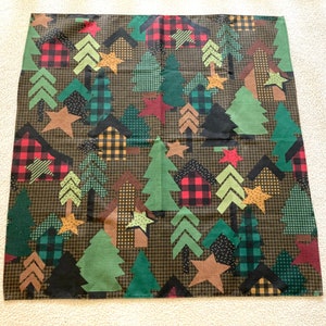 May include: A black fabric with a repeating pattern of green trees, red and black plaid houses, and gold stars. The fabric has a rustic, woodland theme.