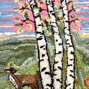 May include: A close-up of a rug with a hand-stitched design of three birch trees with white bark and black markings. The trees are in front of a pink and blue sky with green foliage. A brown deer is in the foreground.