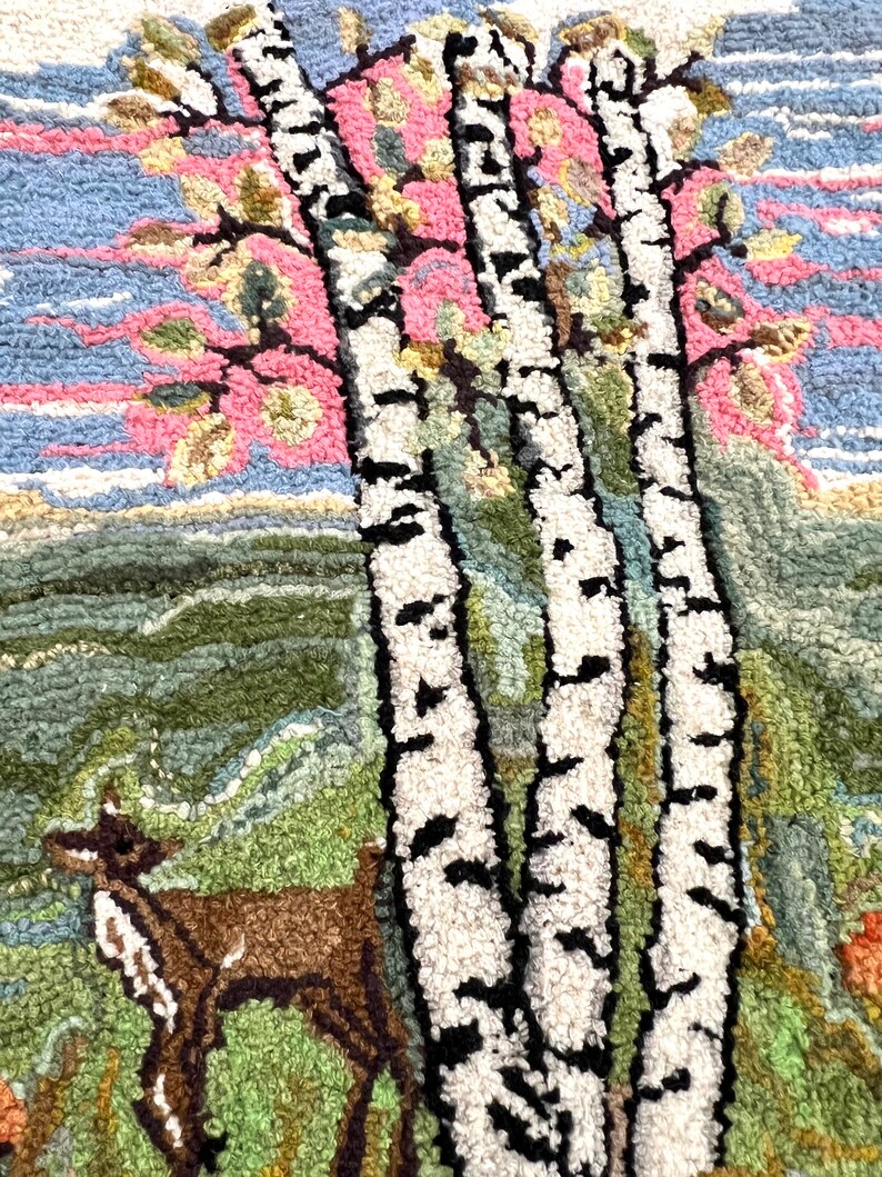 May include: A close-up of a rug with a hand-stitched design of three birch trees with white bark and black markings. The trees are in front of a pink and blue sky with green foliage. A brown deer is in the foreground.