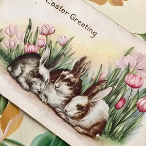 May include: Vintage Easter greeting card featuring three bunnies nestled among pink tulips and green grass. The card has the text "An Easter Greeting" in black script. The background has a soft, pastel color palette.