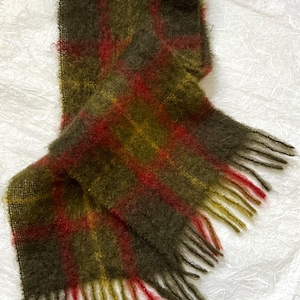 May include: A rectangular scarf with a plaid pattern in shades of olive green, red, and brown. The scarf has a fringed edge and is made of a fuzzy, textured material. The scarf is folded over on itself.