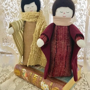 May include: Two handmade fabric dolls with closed eyes and brown hair. The dolls are wearing scarves and long dresses. The doll on the left is wearing a green and white striped dress and a yellow scarf. The doll on the right is wearing a burgundy dress and a tan scarf. The dolls are sitting on top of an old book.