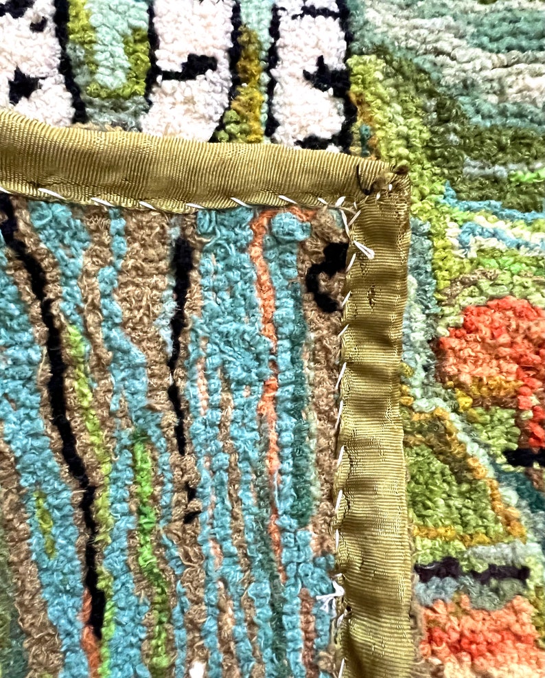 May include: Close-up of a colorful rug with a green, blue, and orange pattern. The rug has a green fabric border with a white stitching detail.