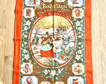 May include: Orange cotton tea towel featuring a colorful illustration of "The Twelve Days of Christmas." The central image depicts a winter scene with figures and a border of holly and flowers. Numbers 1-12 are displayed around the central image.