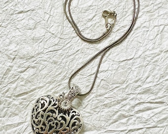 May include: A silver-toned necklace with a heart-shaped pendant. The pendant features an open filigree design and a small, clear gemstone. The necklace chain is a thin, snake chain style, and the clasp is visible.