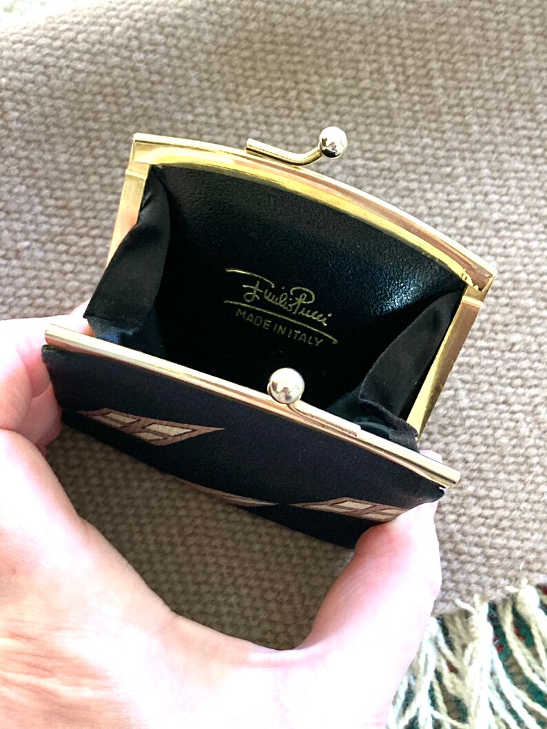 May include: A black and gold coin purse with a geometric pattern. The purse is open and the inside is lined with black fabric. The purse has a gold clasp and the words "Emilio Pucci MADE IN ITALY" are printed on the inside.