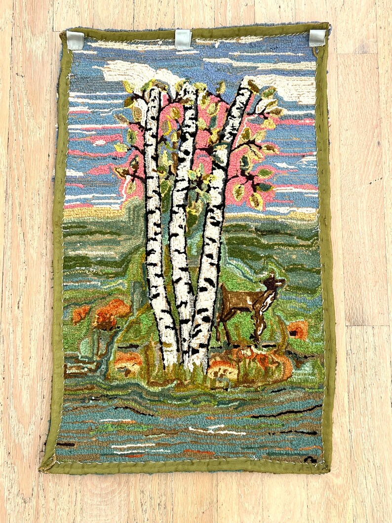 May include: A vintage hooked rug depicting a scene of three birch trees with a deer standing in front of them. The trees are white with black markings and the deer is brown. The background is a blue sky with pink clouds and a green field.