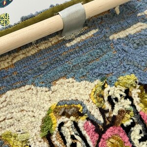 May include: A close-up of a colorful rug with a floral design. The rug is attached to a wooden dowel with a gray fabric loop.