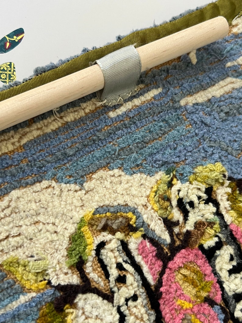 May include: A close-up of a colorful rug with a floral design. The rug is attached to a wooden dowel with a gray fabric loop.