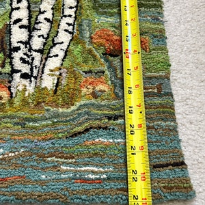 May include: A close-up of a hand-woven rug with a landscape scene featuring birch trees, a river, and mushrooms. The rug is made with a variety of colors, including green, brown, blue, and white. The rug is approximately 23 inches long.