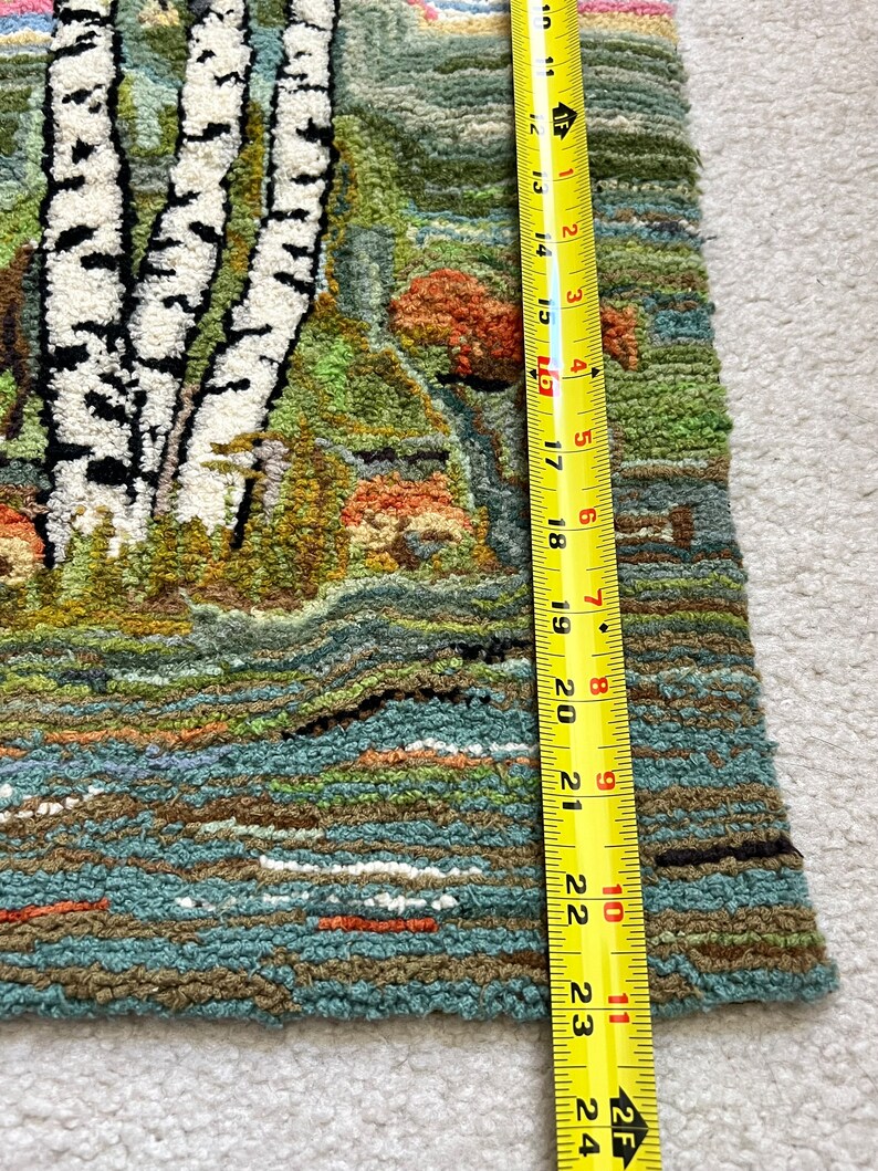May include: A close-up of a hand-woven rug with a landscape scene featuring birch trees, a river, and mushrooms. The rug is made with a variety of colors, including green, brown, blue, and white. The rug is approximately 23 inches long.