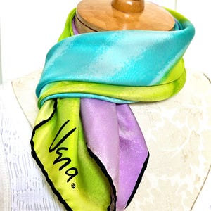 May include: A colorful silk scarf with a vibrant design. The scarf features shades of blue, green, and purple. The scarf has the word "Vana" in black script on a corner. The scarf is draped over a wooden form.