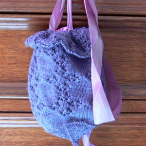 Pode incluir: Uma bolsa de malha roxa com uma parte superior franjada e uma alça de fita rosa. A bolsa está pendurada em um gancho prateado em uma superfície de madeira.