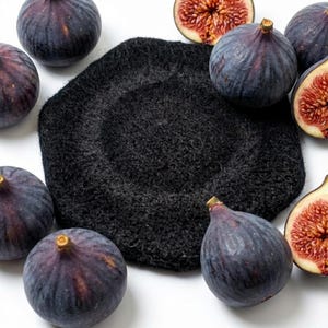 May include: A black beret sits among fresh figs on a white surface. The figs are a deep purple color, with some cut open to reveal the red interior. The beret is a dark, textured fabric.