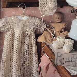 May include: A crocheted baby gown and bonnet set in a cream color. The gown has a delicate lace pattern and a tie at the waist. The bonnet is a simple design with a scalloped edge.