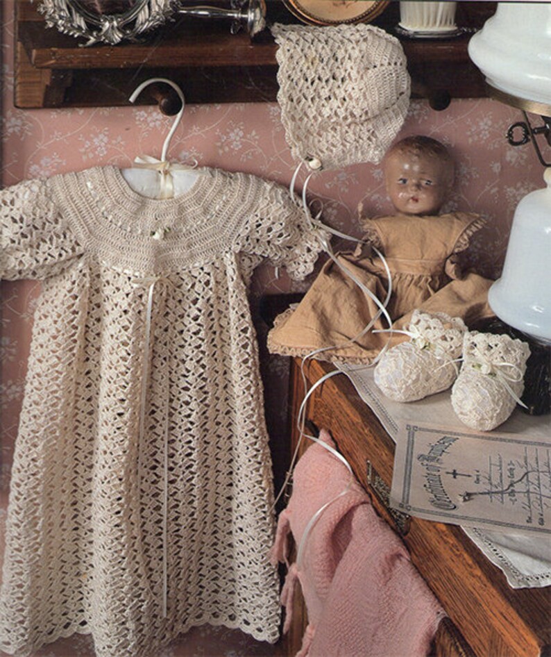 May include: A crocheted baby gown and bonnet set in a cream color. The gown has a delicate lace pattern and a tie at the waist. The bonnet is a simple design with a scalloped edge.