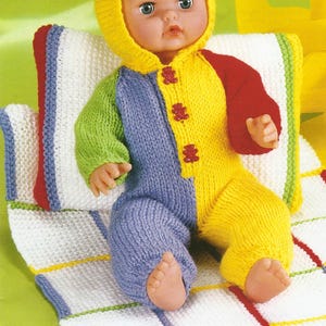 May include: A doll dressed in a knitted jumpsuit with a hood, featuring sections of blue, green, yellow, and red. The doll rests on a white blanket with coloured stripes and a matching pillow, all knitted.