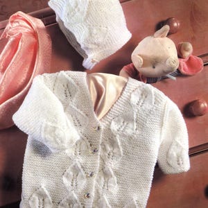 May include: A white knitted baby set, including a cardigan, hat, and a stuffed pig toy. The cardigan features leaf-shaped designs and button closures. The hat has a leaf design. The set is displayed on a wooden surface.