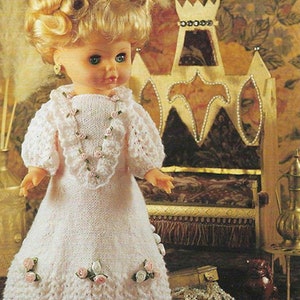 May include: A white knitted doll dress with a lace trim and pink flower accents. The dress is worn by a doll with blonde hair and blue eyes. The doll is sitting on a throne-like chair with a crown on top. The text "LITTLE PRINCESS" is visible in the upper right corner of the image.