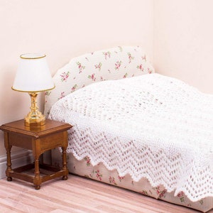 May include: A white crocheted bedspread with a scalloped edge, covering a floral patterned bed with a gold table lamp on a wooden side table.