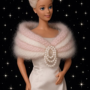 Könnte beinhalten: Puppe in einem weißen Satin-Kleid und einem rosa-weiß gestreiften Schal. Eine Perlenbrosche verziert den Schal. Die Puppe hat blondes Haar und blaue Augen, vor einem schwarzen Hintergrund mit weißen Sternen.