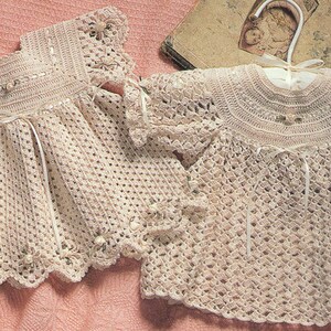 May include: Two crocheted baby dresses in a cream color. The dresses have a delicate lace trim and are made with a fine yarn.