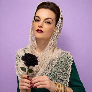 May include: A woman wearing a cream-colored, open-weave shawl and head covering, holding a black rose. She wears a green top and gold bracelets. The background is a solid purple.