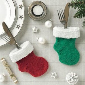 May include: Two knitted Christmas stocking-shaped cutlery holders, one red and one green, with white fur trim. The holders are filled with a fork and knife.