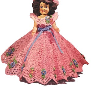 Könnte beinhalten: Vintage Puppe in einem handgefertigten, rosa gehäkelten Kleid und einer Haube. Das Kleid hat einen weiten Rock, der mit blauen und gelben Blumenmotiven verziert ist. Ein blaues Band betont die Taille.