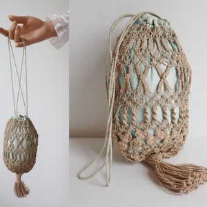 Può includere: Una borsa con coulisse in macramè beige con interno azzurro. La borsa ha una lunga tracolla in corda beige e un nappa abbinata sul fondo. Il design in macramè crea un motivo aperto e tessuto.