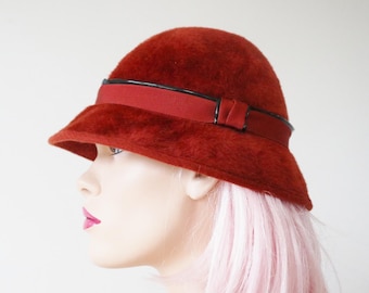 May include: A cloche hat in a deep red hue, featuring a matching ribbon band with a black trim. The hat has a rounded crown and a downward-sloping brim. The hat is a vintage style.