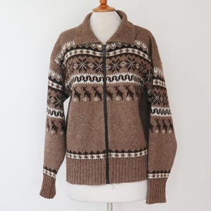 Könnte beinhalten: Ein brauner Reißverschluss-Cardigan mit einem gemusterten Design. Der Cardigan hat einen Kragen und einen schwarzen Reißverschluss. Das Muster umfasst weiße, braune und schwarze geometrische Formen und Rentiere. Er hat gerippte Bündchen und Saum.