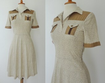May include: A vintage-style dress in beige with a subtle floral pattern. It features a collared neckline, short sleeves, and two front pockets with brown accents. The dress has a fitted bodice and a flared skirt, creating a classic silhouette.