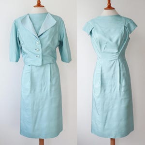 以下が含まれることがあります： マネキンに展示された2つのヴィンテージライトブルーのドレス。1つは半袖ジャケットと膝丈スカートのツーピースセットです。もう1つは、フィットしたボディスと膝丈スカートのノースリーブドレスです。