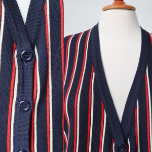 Op de afbeelding: Marineblau vest met verticale rode en witte strepen. De trui met knopen wordt op een mannequin gepresenteerd. De close-up toont de details van de knopen en de gebreide stof.