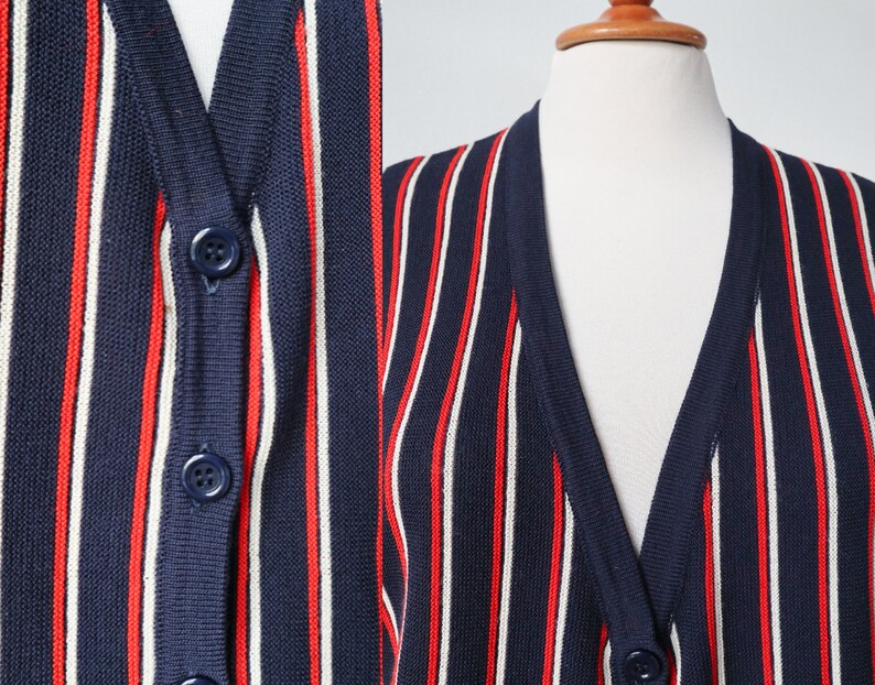 Op de afbeelding: Marineblau vest met verticale rode en witte strepen. De trui met knopen wordt op een mannequin gepresenteerd. De close-up toont de details van de knopen en de gebreide stof.