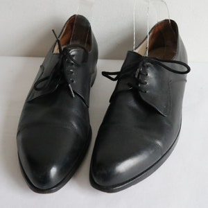 May include: A pair of black leather dress shoes with a classic Oxford design. The shoes have a pointed toe, black laces, and a low heel. The shoes are displayed on a white surface.