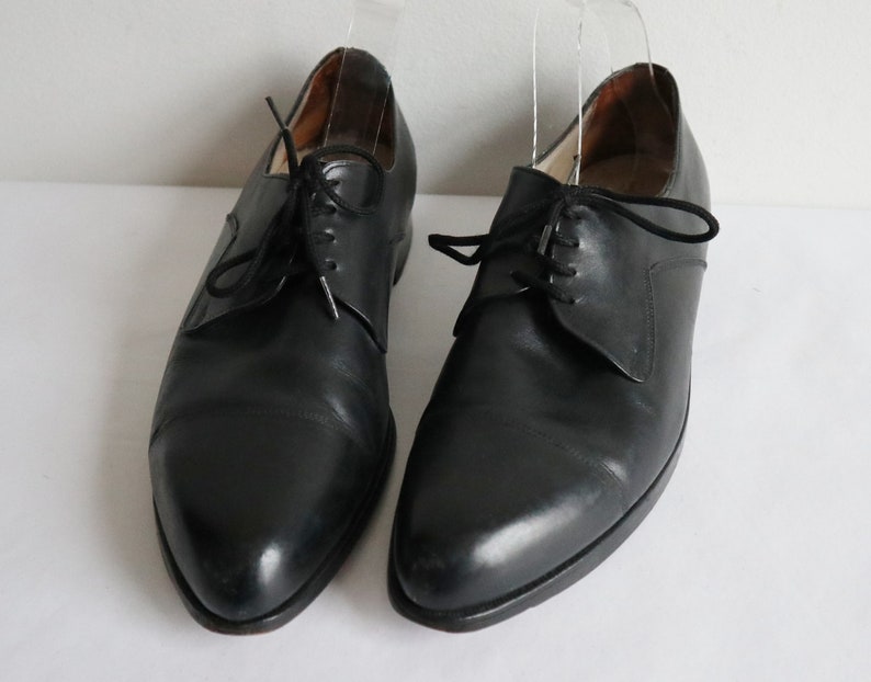 May include: A pair of black leather dress shoes with a classic Oxford design. The shoes have a pointed toe, black laces, and a low heel. The shoes are displayed on a white surface.