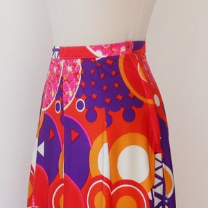 May include: A vibrant, high-waisted skirt featuring a bold, geometric pattern. The design incorporates large circles, triangles, and other shapes in shades of red, orange, purple, and white. The skirt has a button closure and a pleated design.